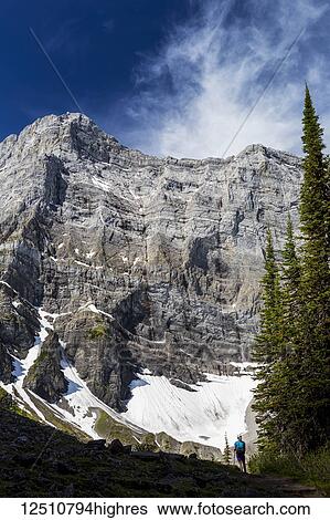 Stock Photograph - Female hiker on ridge looking at large mountain with cloud and blue sky; Kananaskis Country, Alberta, Canada. Fotosearch