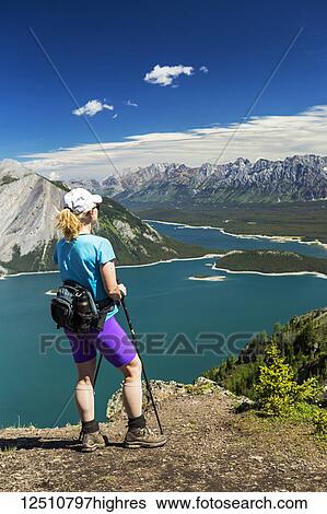 Female hiker on ridge top overlooking a colourful alpine lake and mountain range in the distance with blue sky and clouds; Kananaskis Country, Alberta, Canada View Large Photo Image Stock Photograph - Female hiker on ridge top overlooking a colourful alpine lake and mountain range in the distance with blue sky and clouds; Kananaskis Country, Alberta, Canada. Fotosearch