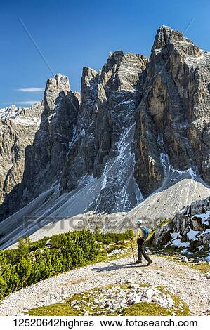 Stock Photograph - Female hiker on trail with valley below against a rugged mountain range and blue sky; Sesto, Bolzano, Italy. Fotosearch