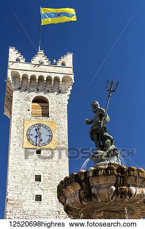 Stock Photograph - Fountain top of Poseidon with trident and large stone clock tower with flag of Trento in the background with blue sky; Trento, Trento, Italy. Fotosearch