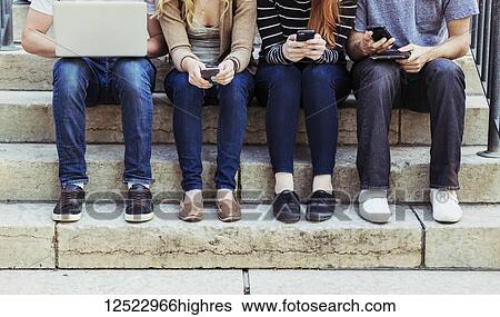 Stock Photograph - Four students sitting in a row on a step using their technology on the university campus, with a view of their feet and legs only; Edmonton, Alberta, Canada. Fotosearch