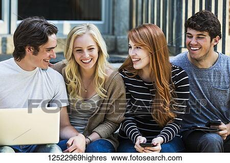 Stock Photograph - Four students sitting in a row on a step using their technology on the university campus and laughing about something funny; Edmonton, Alberta, Canada. Fotosearch
