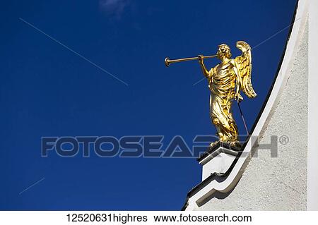 Gold winged angel sculpture with trumpet on top of white church against a blue sky; San Candido, Bolzano, Italy View Large Photo Image Stock Photograph - Gold winged angel sculpture with trumpet on top of white church against a blue sky; San Candido, Bolzano, Italy. Fotosearch