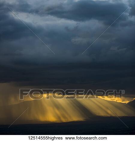 Stock Photograph - Golden sun rays and illuminated rainfall shining through the dark, ominous clouds with mountains in the distance, Volcanoes National Park; Northern Province, Rwanda. Fotosearch