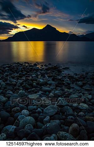 Golden sunlight glowing behind a silhouetted mountain and a tranquil lake with rocky shore in the foreground; Vancouver, British Columbia, Canada View Large Photo Image Stock Photograph - Golden sunlight glowing behind a silhouetted mountain and a tranquil lake with rocky shore in the foreground; Vancouver, British Columbia, Canada. Fotosearch