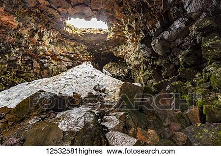 Stock Photograph - Hiking through the lava tube caves near Reykjavik in early summer, a pile of snow has not quite melted yet from the cave. Visitors here walk the path of lava that flowed during an eruption; Iceland. Fotosearch