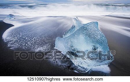 Stock Photograph - Jagged piece of broken ice laying on the sandy shore at the edge of the ocean water; Iceland. Fotosearch