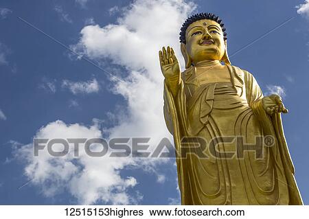 Stock Photograph - Low angle view of a gold Buddha statue against a blue sky with cloud; Ulaanbaatar, Ulaanbaatar, Mongolia. Fotosearch