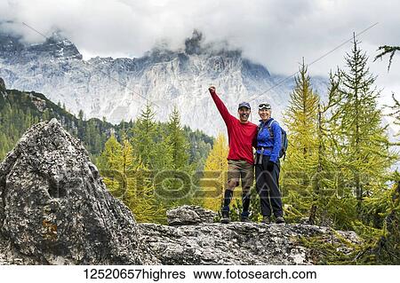 Male and female hikers standing on rock with autumn coloured trees and cloud-covered mountain range in the background; Sesto, Bolzano, Italy View Large Photo Image Stock Photograph - Male and female hikers standing on rock with autumn coloured trees and cloud-covered mountain range in the background; Sesto, Bolzano, Italy. Fotosearch