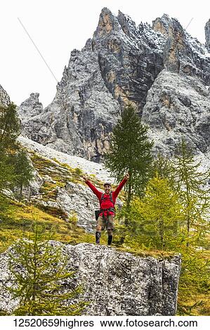 Stock Photograph - Male hiker with arms raised on top of large rock with rocky slope and mountain peak; Sesto, Bolzano, Italy. Fotosearch