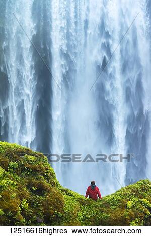 Stock Photograph - Man sitting on a mossy rock at base of Skogafoss waterfall; Iceland. Fotosearch