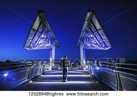 Stock Photograph - Man walking on Terwillegar Park bridge at dusk; Edmonton, Alberta, Canada. Fotosearch