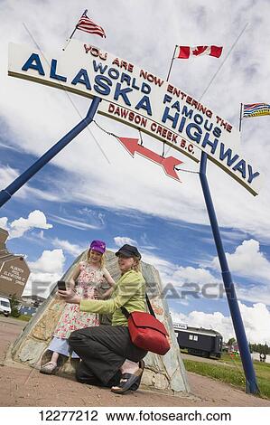 Stock Image - Mother and daughter take a selfie underneath the World Famous Alaska Highway sign, Dawson Creek, British Columbia, Canada. Fotosearch