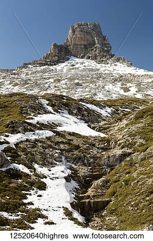 Stock Photograph - Mountain peak above a snow-covered slope with small creek flowing down and blue sky; Sesto, Bolzano, Italy. Fotosearch