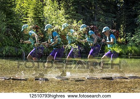 Stock Photograph - Multiple exposure of a female hiker with trekking poles leaps from rock to rock in a shallow stream and splashes water with her boots; Calgary, Alberta, Canada. Fotosearch