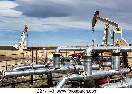 Stock Image - Pipelines with two pumpjacks in the background with cloud cover and blue sky; Alberta, Canada. Fotosearch