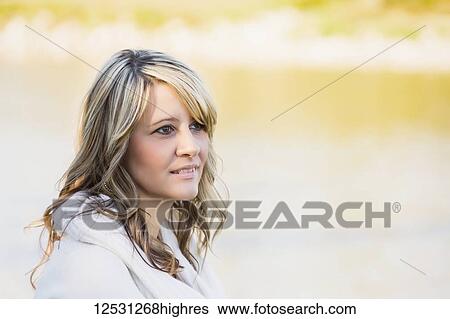 Stock Photograph - Portrait of a beautiful mature woman with blond hair in a city park with autumn colours in the background reflected on tranquil water; Edmonton, Alberta, Canada. Fotosearch