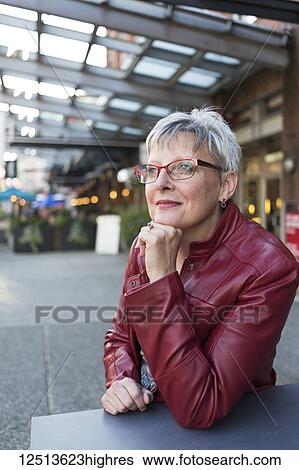 Stock Photograph - Portrait of a mature woman in a red leather jacket sitting at a table outside a shop with her chin resting in her hand; Vancouver, British Columbia, Canada. Fotosearch