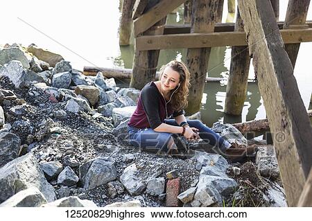 Stock Photograph - Portrait of a young woman sitting on rocks under a bridge by water listening to music. Fotosearch