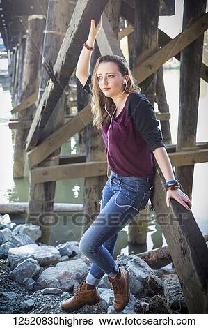 Stock Photograph - Portrait of a young woman standing on rocks under a bridge by water listening to music; New Westminster, British Columbia, Canada. Fotosearch