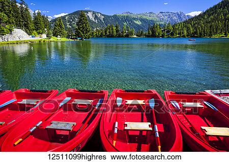 Stock Photograph - Red boats lined up at Champex Lake under blue sky with a mountain range in the background; Champex, Valais, Switzerland. Fotosearch