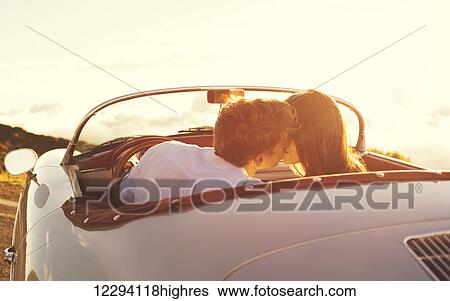 Stock Photograph - Romantic Young Attractive Couple Watching The Sunset And Kissing In Classic Vintage Sports Car. Fotosearch