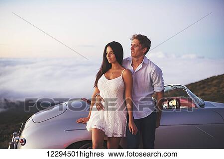 Stock Photograph - Romantic Young Attractive Couple Watching The Sunset With Classic Vintage Sports Car. Fotosearch