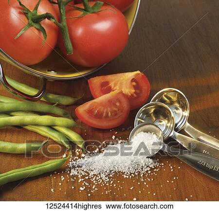 Stock Photograph - Salt from a measuring spoon with fresh green beans and tomatoes on a wooden surface. Fotosearch