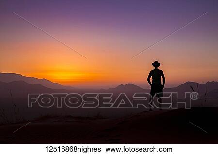 Silhouette of a man wearing a cowboy hat stands looking out at the glowing landscape and sky at sunset; Sossusvlei, Hardap Region, Namibia View Large Photo Image Stock Photograph - Silhouette of a man wearing a cowboy hat stands looking out at the glowing landscape and sky at sunset; Sossusvlei, Hardap Region, Namibia. Fotosearch