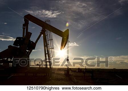 Stock Photograph - Silhouette of a pumpjack backlit by the setting sun on the Alberta Prairies with blue sky and cloud, West of Airdrie; Alberta, Canada. Fotosearch
