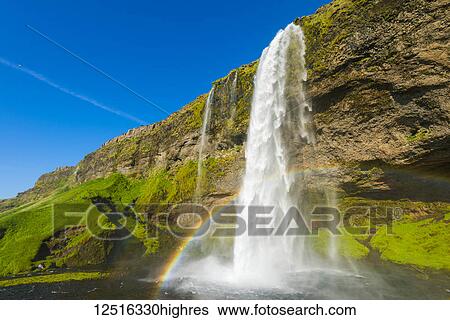 Stock Photograph - Skogafoss waterfall with blue sky and a rainbow in the mist; Iceland. Fotosearch