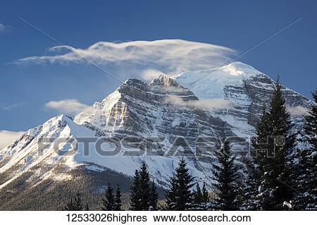 Stock Photograph - Snow-covered mountain with wispy cloud formation on peak and blue sky, Banff National Park; Lake Louise, Alberta, Canada. Fotosearch