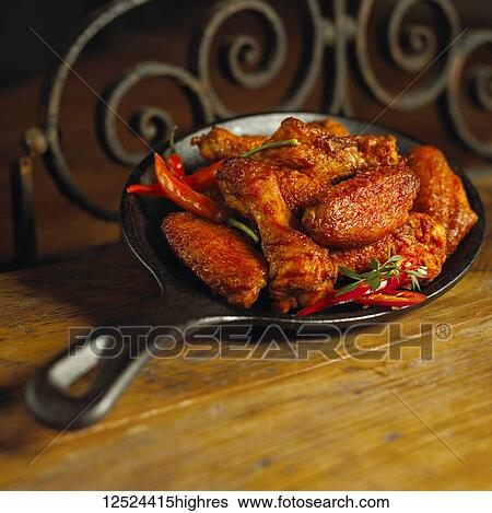 Stock Photograph - Spicy chicken wings and peppers in a cast iron pan on a wooden counter. Fotosearch