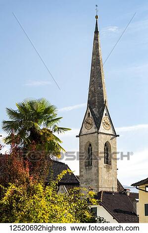 Stock Photograph - Stone church clock tower in alpine village with palm tree and blue sky; Caldaro, Bolzano, Italy. Fotosearch