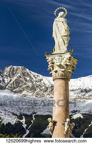 Stock Photograph - Stone statue with halo on top of column with snow-covered mountain range and blue sky in the background; Innsbruck, Tyrol, Austria. Fotosearch