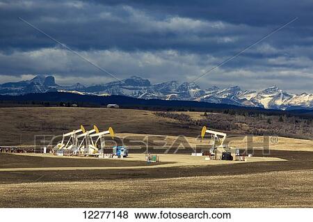 Sun lit pumpjacks in a field with dark clouds in the sky, shadowed rolling hills and mountains in the background; Alberta, Canada View Large Photo Image Stock Photo - Sun lit pumpjacks in a field with dark clouds in the sky, shadowed rolling hills and mountains in the background; Alberta, Canada. Fotosearch