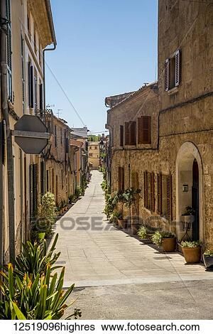 Stock Photograph - Sunny narrow street lined with houses and decorative plants with a blue sky; Alcudia, Mallorca, Balearic Islands, Spain. Fotosearch