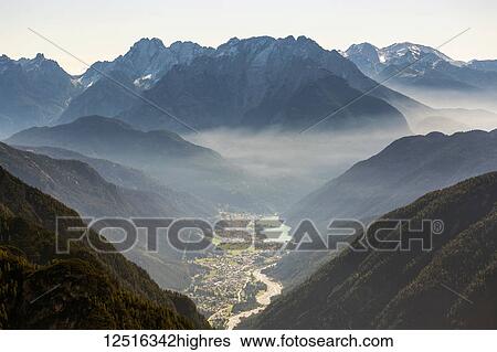 Stock Photograph - Sunrise with illuminated fog laying through the mountain range and over a valley; Sesto, Bolzano, Italy. Fotosearch