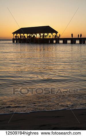 Stock Photograph - Sunset over ocean and silhouette of tourists on Hanalei Pier, Hanalei Bay; Hanalei, Kauai, Hawaii, United States of America. Fotosearch