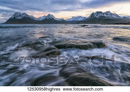 Stock Photograph - Sunset over the mountains of the Strandir Coast, Iceland as the surf pounds the shoreline; West Fjords, Iceland. Fotosearch