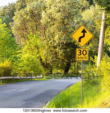Stock Photograph - Sunset Road on the way to the Lighthouse on Mayne Island, a country road with a road sign indicating a curve and signal to a lower speed; Mayne Island, British Columbia, Canada. Fotosearch