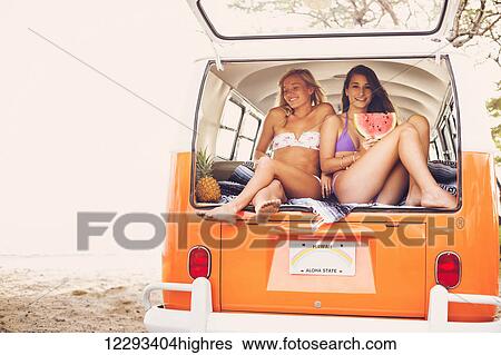 Stock Photograph - Surfer Girls Beach Lifestyle, Friends Hanging Out Eating Watermelon In The Back Of Classic Vintage Surf Van On The Beach At Sunset. Fotosearch