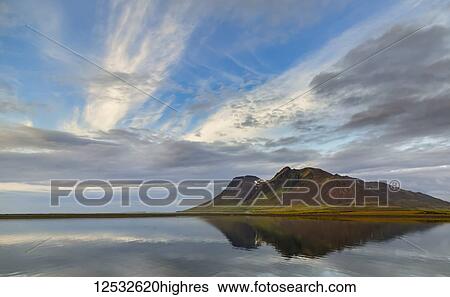 Stock Photograph - The rich volcanic earth creates bold colours on the mountain against the soft blue ocean and sky reflection in Western Iceland; Iceland. Fotosearch