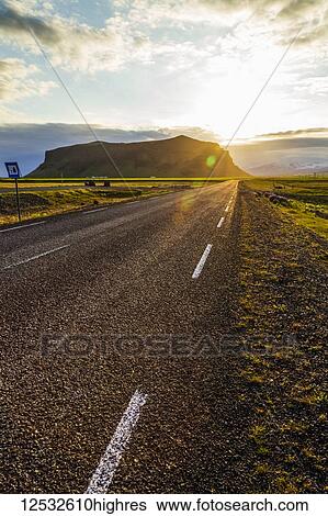 Stock Photograph - The sun sets behind the hills with a paved highway road leading into the sunset; Iceland. Fotosearch