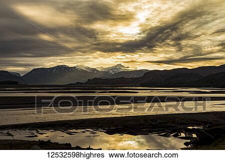 Stock Photograph - The sun sets late in the evening near summer solstice and silhouettes the mountains and river on this remote landscape; Iceland. Fotosearch