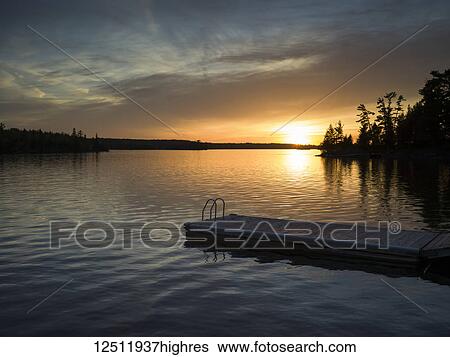 The sun setting over a tranquil lake and silhouetted trees along the shoreline with a dock and ladder in the foreground; Lake of the Woods, Ontario, Canada View Large Photo Image Stock Photograph - The sun setting over a tranquil lake and silhouetted trees along the shoreline with a dock and ladder in the foreground; Lake of the Woods, Ontario, Canada. Fotosearch