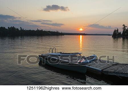 Stock Photograph - The sun setting over a tranquil lake with a dock, canoe and paddle boards in the foreground; Lake of the Woods, Ontario, Canada. Fotosearch