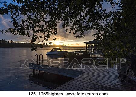 The sun setting over a tranquil lake with a dock and boat in the foreground; Lake of the Woods, Ontario, Canada View Large Photo Image Stock Photograph - The sun setting over a tranquil lake with a dock and boat in the foreground; Lake of the Woods, Ontario, Canada. Fotosearch