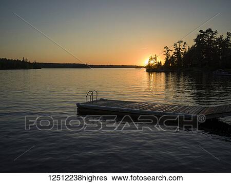 Stock Photograph - The sun setting over Lake of the Woods with a dock in the foreground; Lake of the Woods, Ontario, Canada. Fotosearch
