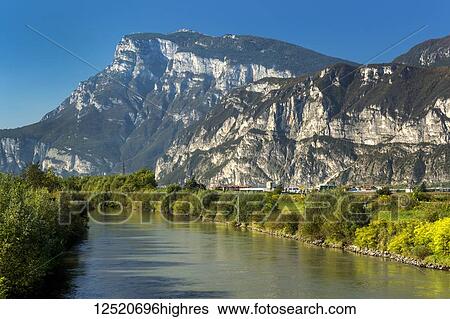 Tranquil river lined with trees in green foliage and rugged mountains in the background with blue sky; Trento, Trento, Italy View Large Photo Image Stock Photograph - Tranquil river lined with trees in green foliage and rugged mountains in the background with blue sky; Trento, Trento, Italy. Fotosearch